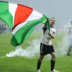 Piłkarz z flagą świętuje zwycięstwo. Czy to on wie, kto ma najwięcej mistrzostw Polski? Dym z rac otacza murawę.