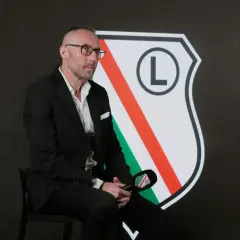 Mężczyzna w garniturze, z charakterystyczną fryzurą i okularami, siedzi przed herbem Legii. To nowy trener Legii Warszawa, gotowy do rozmowy.