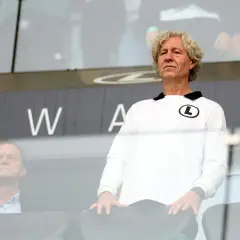 Mężczyzna w białej bluzie z logo Legii, wpatrzony w dal, jakby analizował najnowsze pogłoski transferowe Legia. Obok siedzi inny mężczyzna.