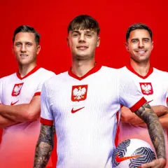 Trzech uśmiechniętych piłkarzy reprezentacji Polski w białych koszulkach z godłem orła. Jeden z nich trzyma piłkę.