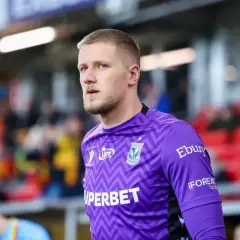 Bartosz Mrozek, bramkarz w fioletowym stroju z wzorem zygzaka, patrzy w bok na tle trybun.
