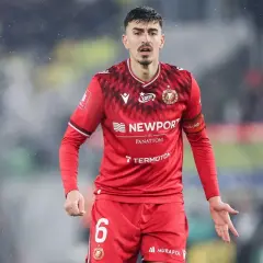 Piłkarz w czerwonej koszulce z numerem 6, z opaską na ramieniu z napisem "Widzew Łódź". W tle widać fragment stadionu, być może podczas meczu z Cracovią.