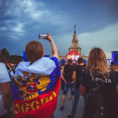 Młody kibic z flagą Rosji, wspierający klub piłkarski z Moskwy, filmuje telefonem wydarzenie na tle chmur i wieżowca.