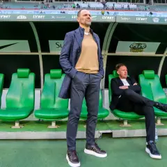 Trener Śląska Wrocław stoi na tle zielonych siedzeń. Obok siedzi inny mężczyzna.