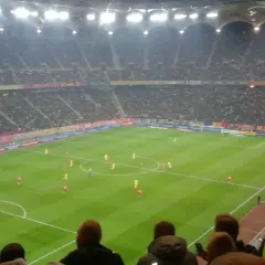 Mecz piłki nożnej na stadionie, tłumy kibiców, flagi Polski i Rumunii.