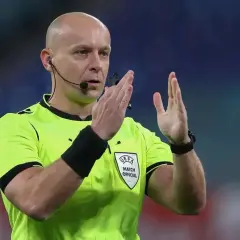 Sędzia piłkarski w żółtej koszulce z emblematem UEFA, z mikrofonem przy uchu, pokazuje gest rękami. Ciekawe, ile zarabia sędzia na takim meczu.