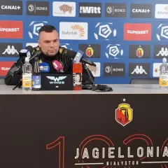 Mężczyzna w czarnym stroju siedzi przy stole konferencyjnym, na którym stoją napoje. Tło zdobią logotypy sponsorów, w tym Jagiellonia Białystok, co sugeruje kontekst związany z jagiellonia transfery.