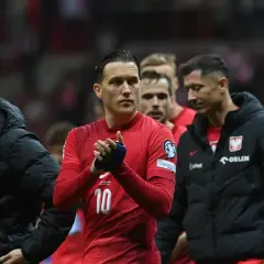 Piłkarze reprezentacji Polski, w tym Lewandowski, w czerwonych strojach i czarnych kurtkach, oklaskują kibiców. Duma z barw narodowych.
