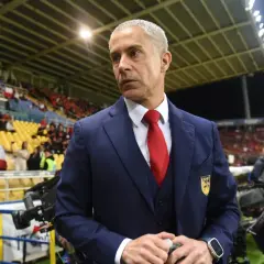 Sylvinho, w granatowym garniturze i czerwonym krawacie, stoi na tle trybun stadionu, gdzie kibice trzymają flagę Chile.