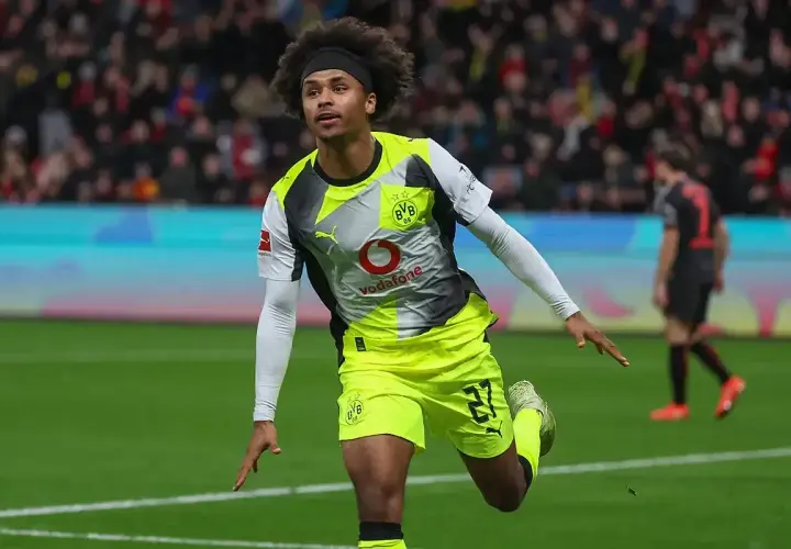 Piłkarz Adeyemi w żółto-szarej koszulce BVB z numerem 27, z afro na głowie, świętuje bramkę na boisku.