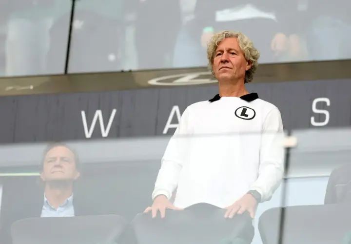 Mężczyzna w białej bluzie z logo Legii, wpatrzony w dal, jakby analizował najnowsze pogłoski transferowe Legia. Obok siedzi inny mężczyzna.