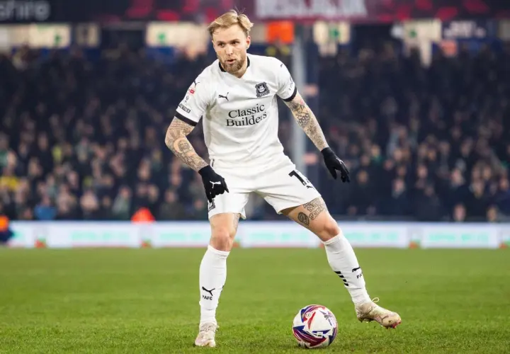Piłkarz w białej koszulce z tatuażami na rękach i nogach, gdzie gra puchacz, przygotowuje się do strzału na zielonej murawie stadionu.
