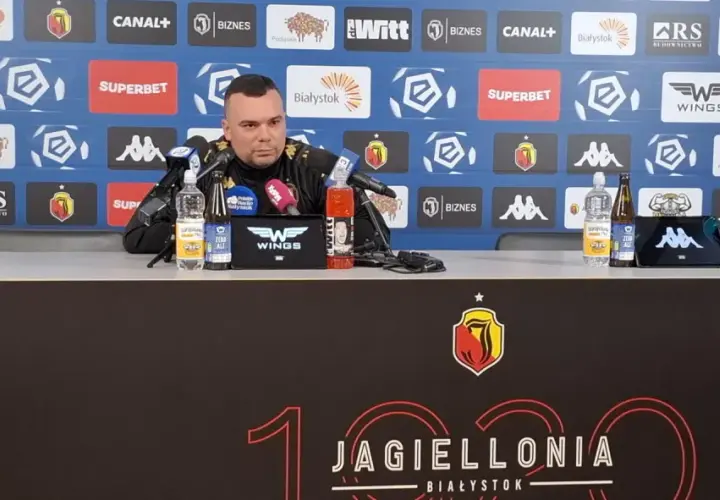 Mężczyzna w czarnym stroju siedzi przy stole konferencyjnym, na którym stoją napoje. Tło zdobią logotypy sponsorów, w tym Jagiellonia Białystok, co sugeruje kontekst związany z jagiellonia transfery.
