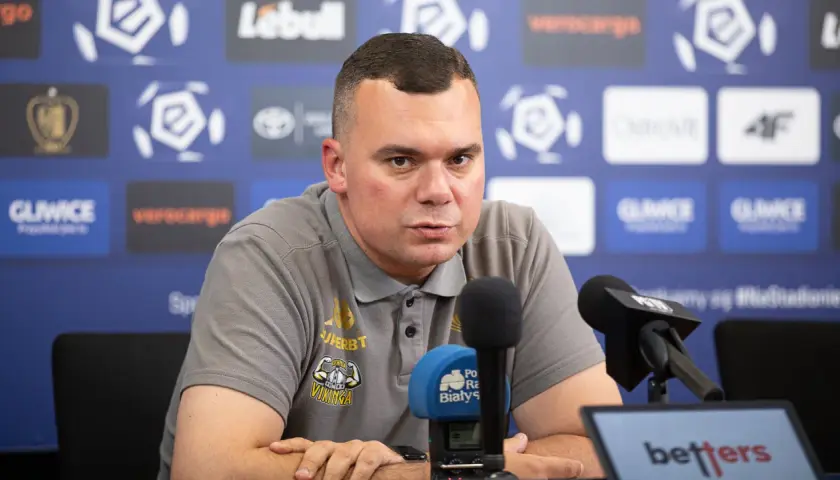 Trener Jagiellonii w szarej koszulce z logo klubu, siedzi przy mikrofonach podczas konferencji prasowej.