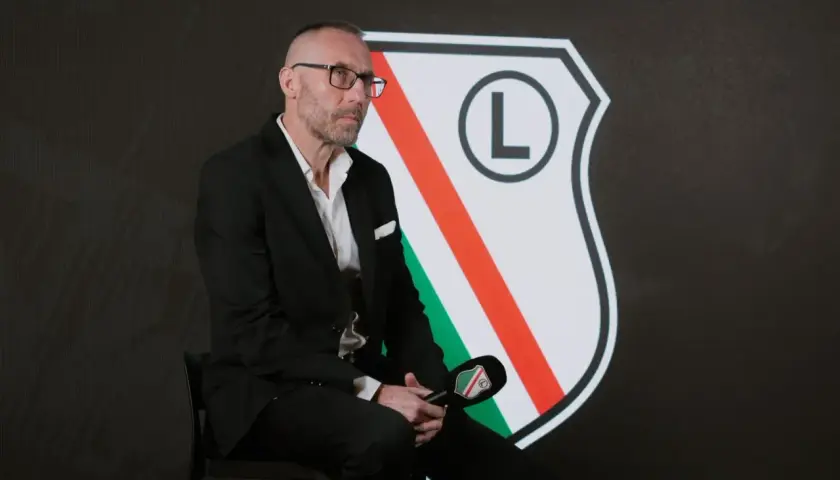 Mężczyzna w garniturze, z charakterystyczną fryzurą i okularami, siedzi przed herbem Legii. To nowy trener Legii Warszawa, gotowy do rozmowy.