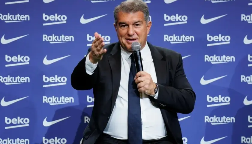Prezes Barcelony, Joan Laporta, przemawia do mikrofonu na tle niebieskiej ścianki z logo klubu i sponsorów.