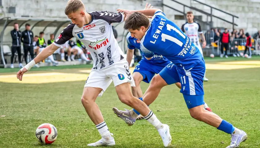 Piłkarze walczą o piłkę na boisku. Jeden w białej koszulce, drugi w niebieskiej z numerem 11. To ważny mecz, który może wpłynąć na 3. liga tabelę.