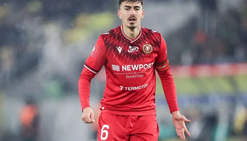 Piłkarz w czerwonej koszulce z numerem 6, z opaską na ramieniu z napisem "Widzew Łódź". W tle widać fragment stadionu, być może podczas meczu z Cracovią.