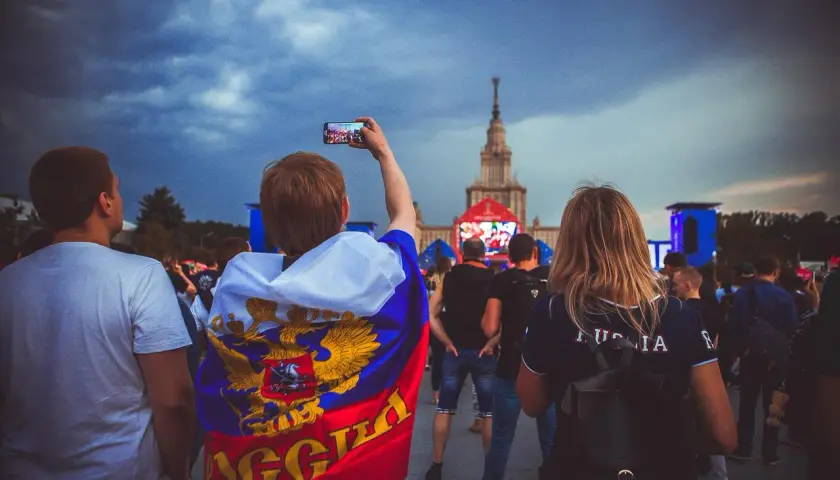Młody kibic z flagą Rosji, wspierający klub piłkarski z Moskwy, filmuje telefonem wydarzenie na tle chmur i wieżowca.