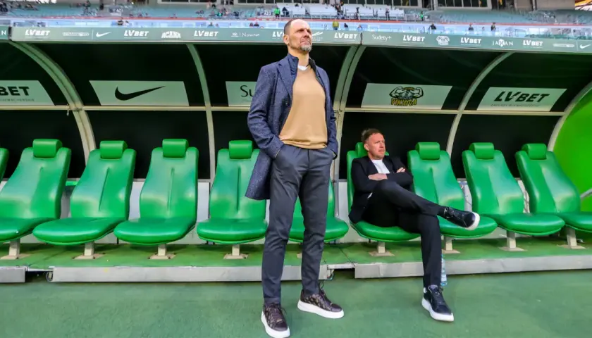 Trener Śląska Wrocław stoi na tle zielonych siedzeń. Obok siedzi inny mężczyzna.