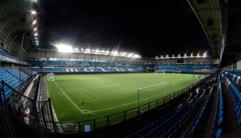 Piłkarski stadion Molde nocą, oświetlony jaskrawymi światłami, z pustymi niebieskimi trybunami i zieloną murawą.