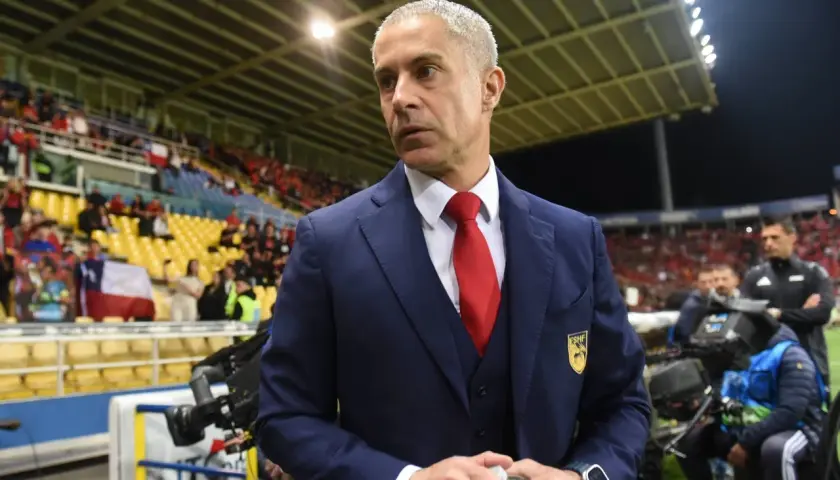 Sylvinho, w granatowym garniturze i czerwonym krawacie, stoi na tle trybun stadionu, gdzie kibice trzymają flagę Chile.