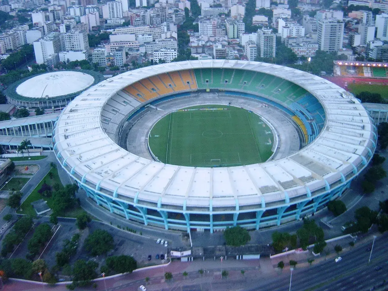 Widok z lotu ptaka na stadion Maracanã z zieloną murawą i kolorowymi trybunami, otoczony miejską zabudową.