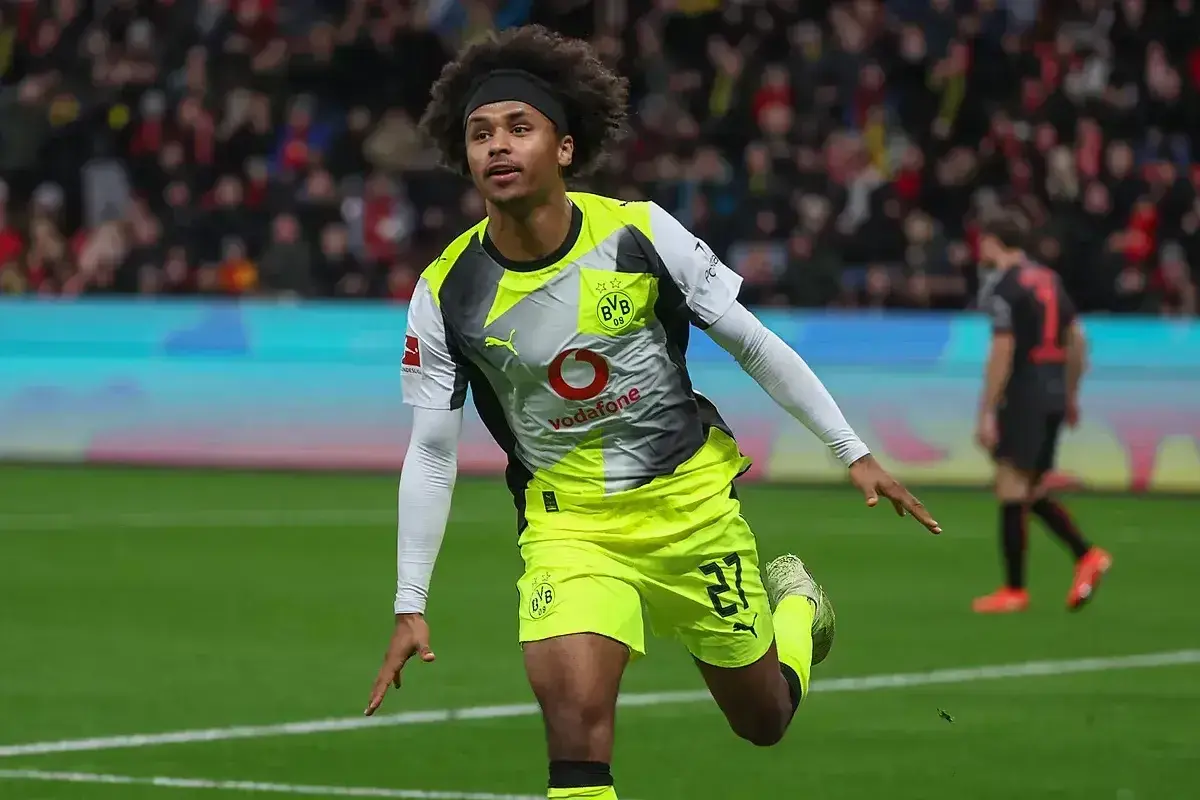 Piłkarz Adeyemi w żółto-szarej koszulce BVB z numerem 27, z afro na głowie, świętuje bramkę na boisku.