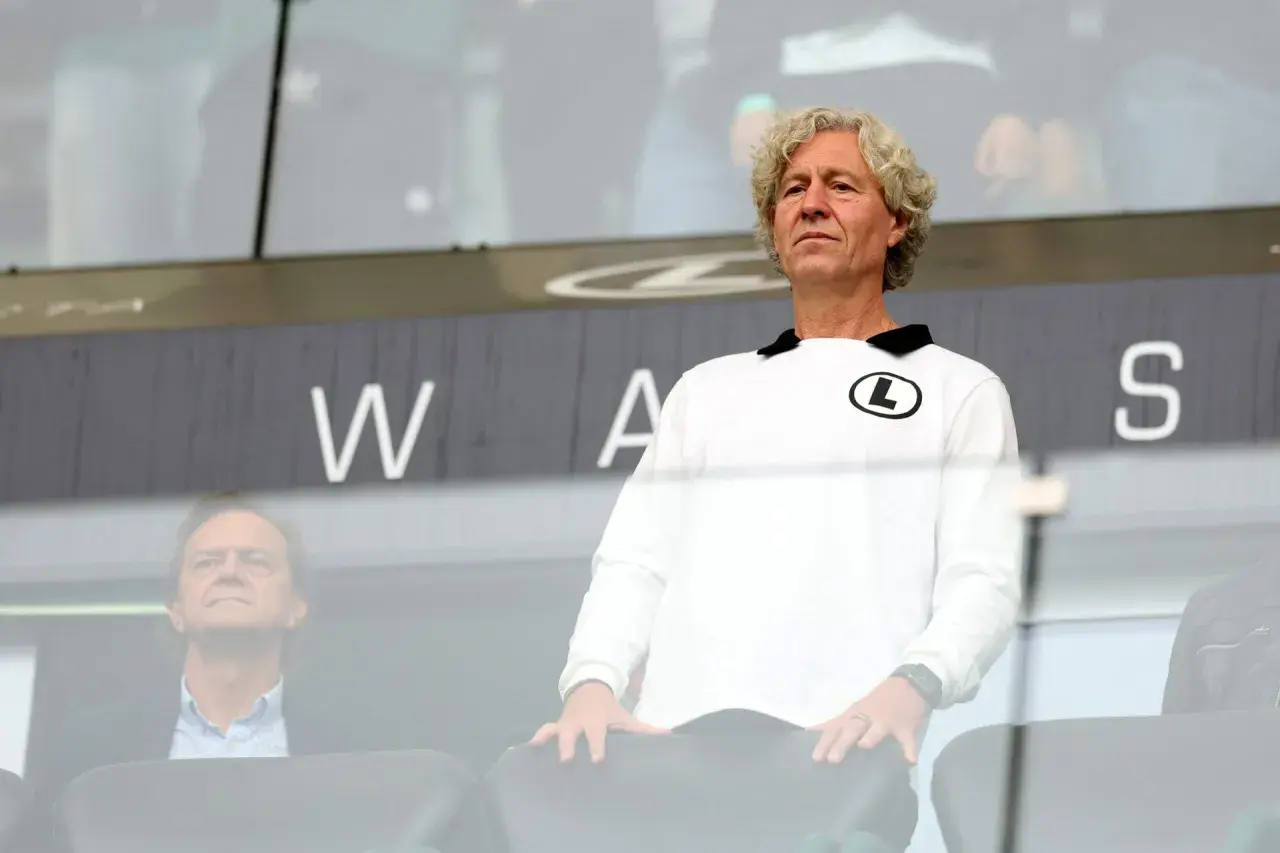 Mężczyzna w białej bluzie z logo Legii, wpatrzony w dal, jakby analizował najnowsze pogłoski transferowe Legia. Obok siedzi inny mężczyzna.