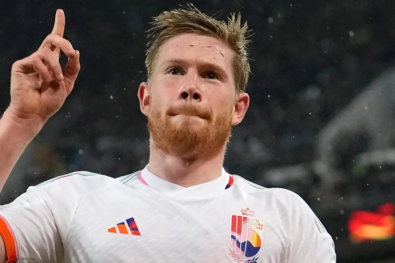 Kevin De Bruyne w białej koszulce z godłem Belgii, unosi palec wskazujący w geście triumfu.