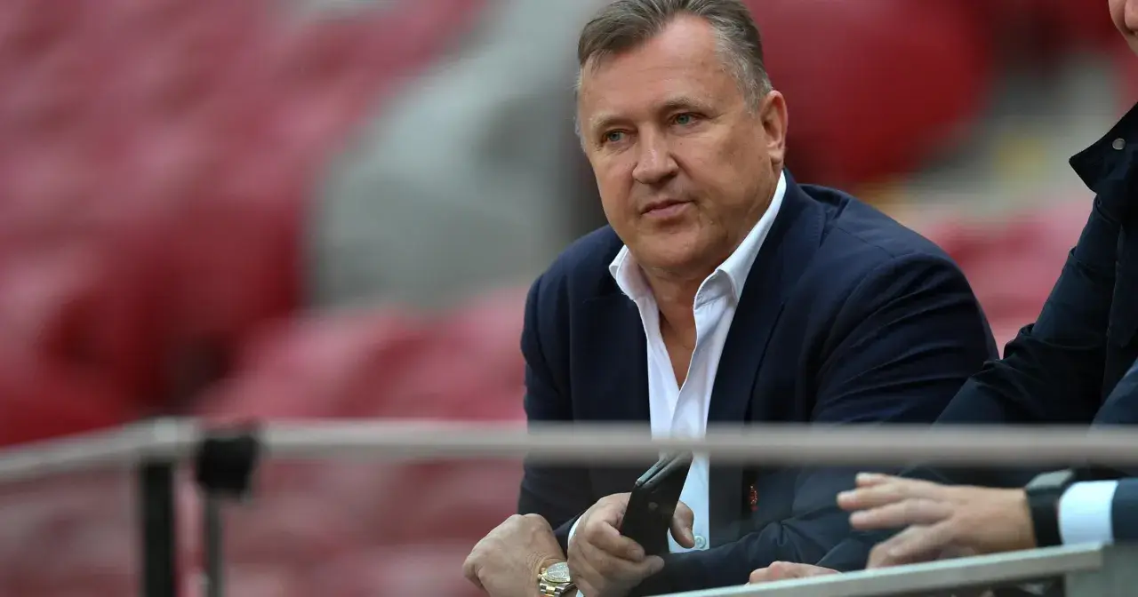 Prezes PZPN w garniturze, z telefonem w dłoni, siedzi na trybunach stadionu.