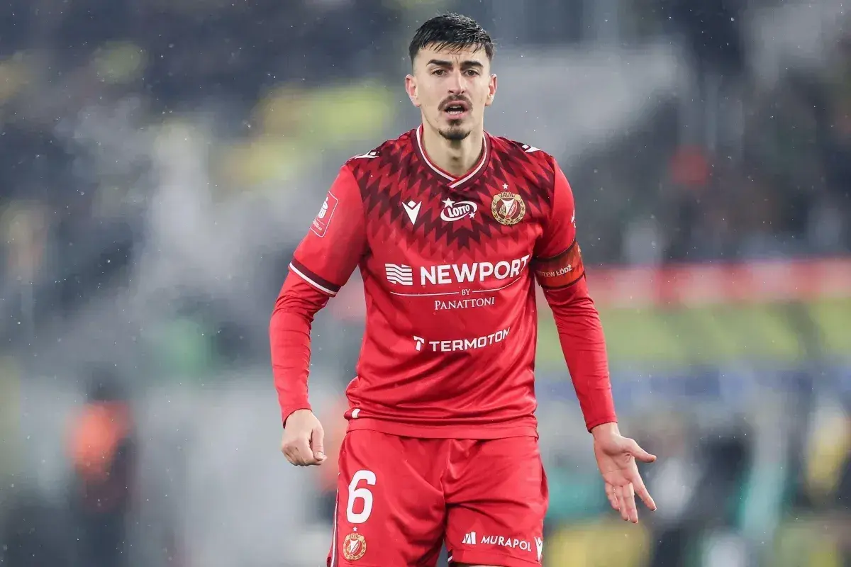 Piłkarz w czerwonej koszulce z numerem 6, z opaską na ramieniu z napisem "Widzew Łódź". W tle widać fragment stadionu, być może podczas meczu z Cracovią.