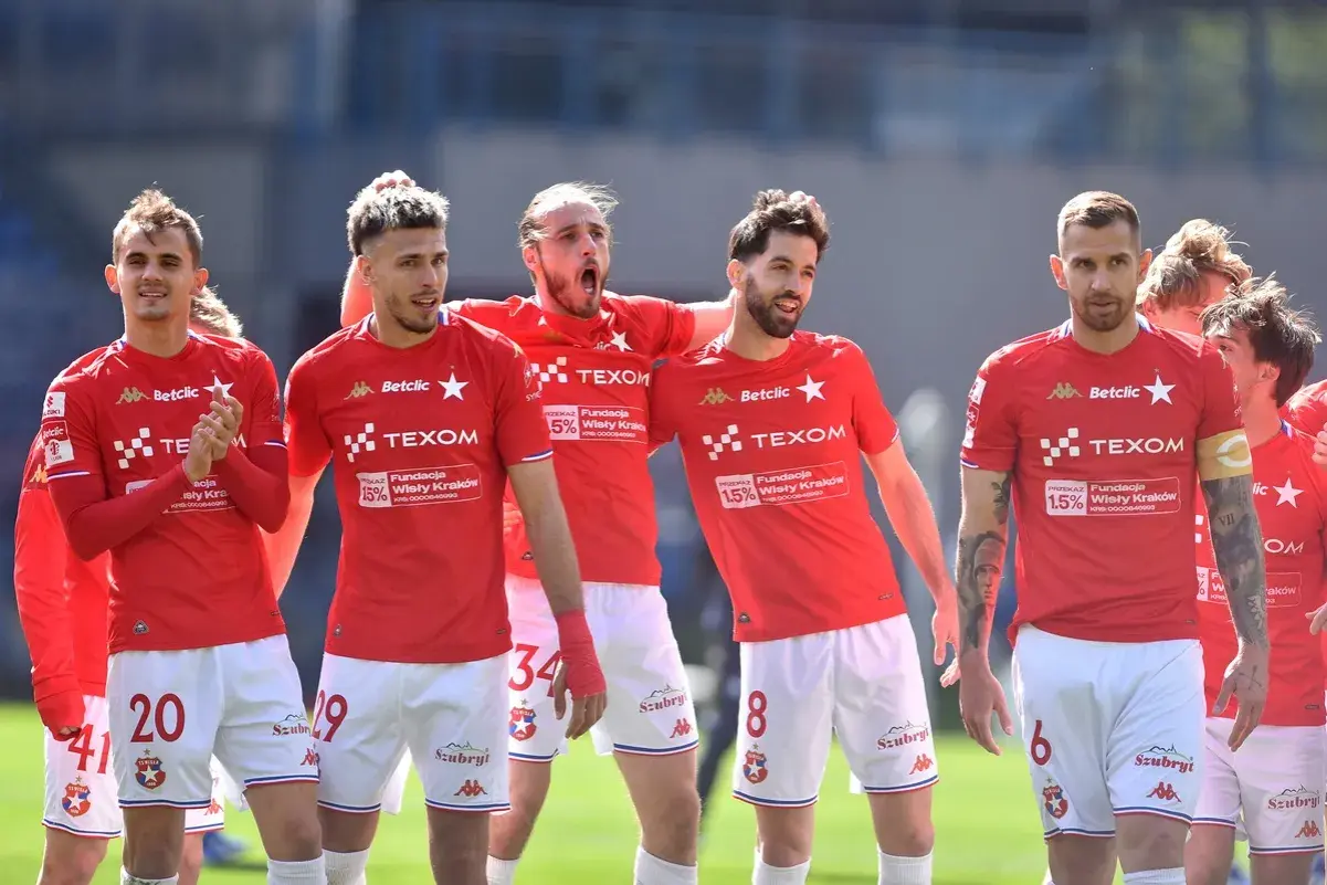 Piłkarze Wisły Kraków w czerwonych strojach, dyskutują o potencjalnych transferach.