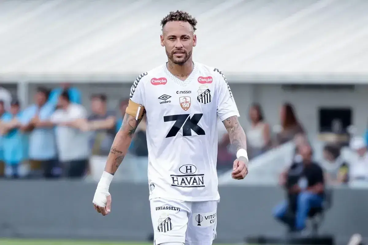 Neymar w białej koszulce Santos, z opaską na ramieniu, na boisku. To właśnie tam gra Neymar, kiedyś gwiazda tego klubu.