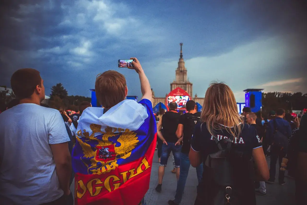 Młody kibic z flagą Rosji, wspierający klub piłkarski z Moskwy, filmuje telefonem wydarzenie na tle chmur i wieżowca.