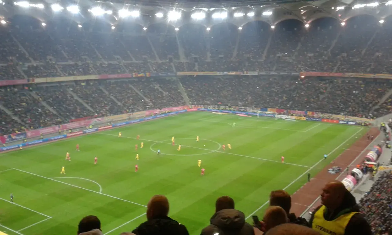 Mecz piłki nożnej na stadionie, tłumy kibiców, flagi Polski i Rumunii.