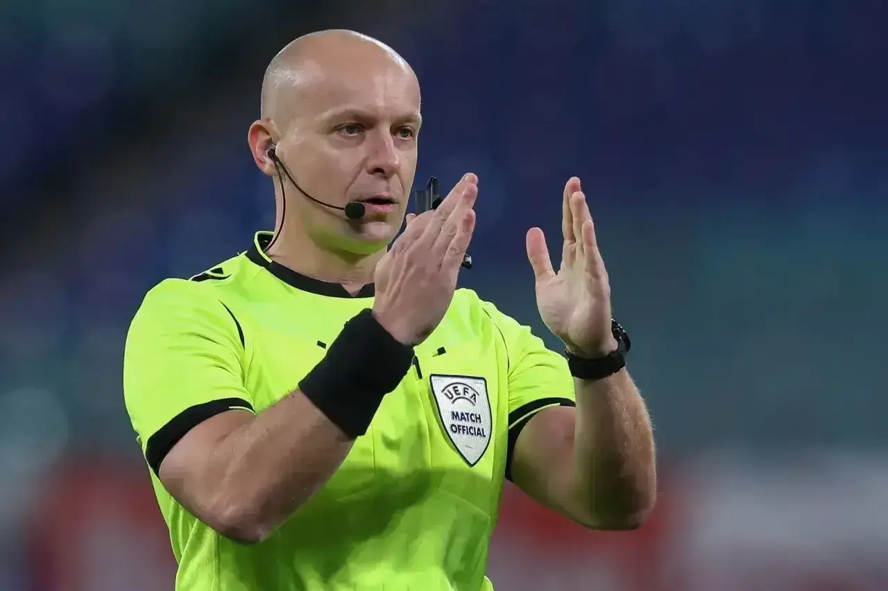 Sędzia piłkarski w żółtej koszulce z emblematem UEFA, z mikrofonem przy uchu, pokazuje gest rękami. Ciekawe, ile zarabia sędzia na takim meczu.
