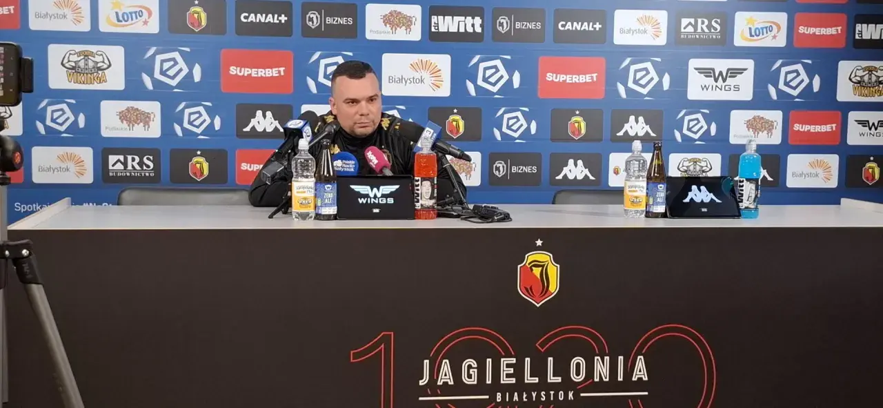 Mężczyzna w czarnym stroju siedzi przy stole konferencyjnym, na którym stoją napoje. Tło zdobią logotypy sponsorów, w tym Jagiellonia Białystok, co sugeruje kontekst związany z jagiellonia transfery.
