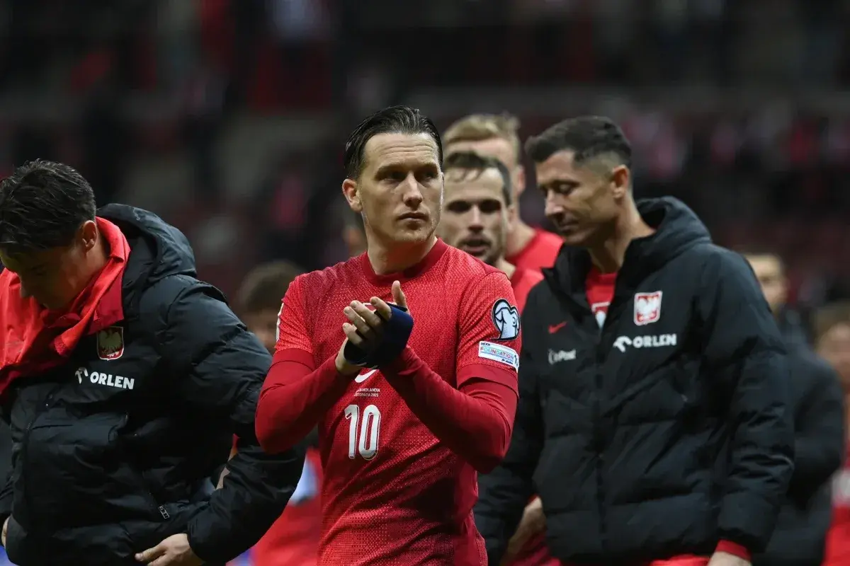 Piłkarze reprezentacji Polski, w tym Lewandowski, w czerwonych strojach i czarnych kurtkach, oklaskują kibiców. Duma z barw narodowych.