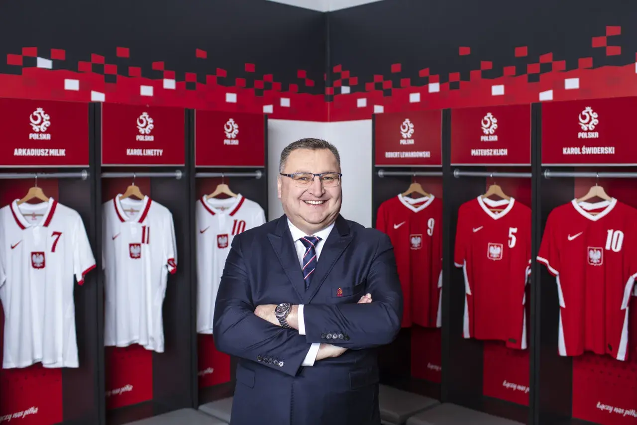 Uśmiechnięty mężczyzna w garniturze stoi przed szatnią reprezentacji Polski. Widać tam koszulki z nazwiskami znanych piłkarzy, tworząc zgrupowanie reprezentacji Polski.