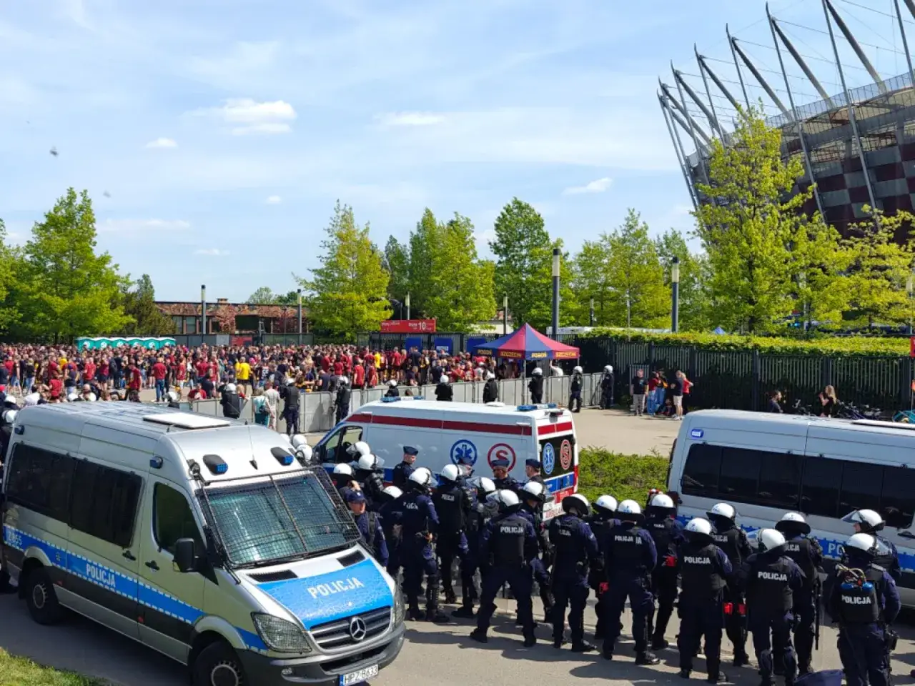 Policja pilnuje porządku przed stadionem, gdzie zebrał się tłum kibiców. Widać radiowozy i funkcjonariuszy w pełnym rynsztunku, gotowych do interwencji, gdyby pojawiły się pseudokibice.