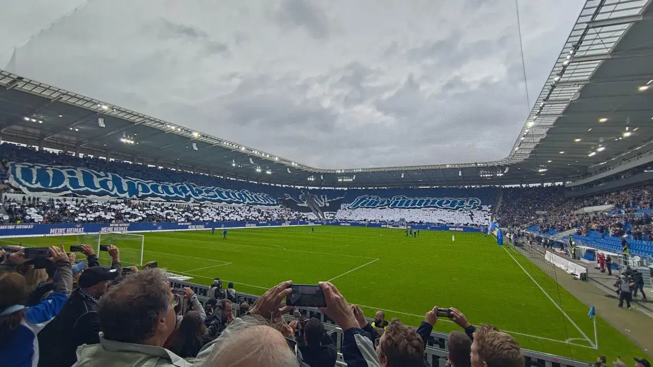 Trybuny stadionu wypełnione kibicami Karlsruher SC. Fani tworzą ogromną mozaikę z hasłami i sylwetką zawodnika.