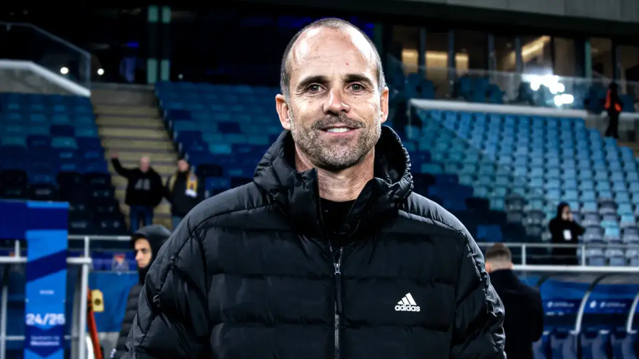 Uśmiechnięty trener Radomiaka w czarnej kurtce z logo Adidas, na tle trybun stadionu.