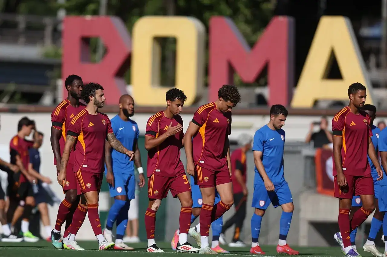 Piłkarze AS Roma w bordowych strojach idą obok zawodników w niebieskich koszulkach. W tle wielki napis ROMA.