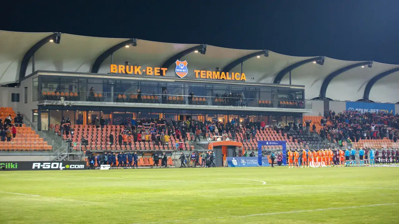 Stadion Bruk-Bet Termalica przed meczem z Jagiellonią Białystok. Piłkarze w pomarańczowych strojach stoją na murawie.