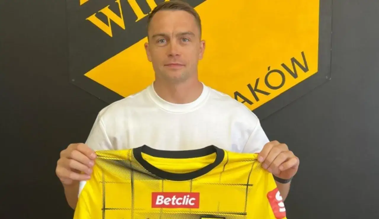 Piłkarz w białej koszulce prezentuje żółtą koszulkę z logo Betclic. Za nim logo klubu z napisem 