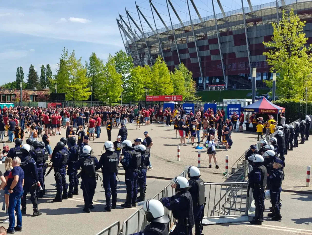 Tłum kibiców przed stadionem, policja pilnuje porządku. Niektórzy pseudokibice wyglądają na zdenerwowanych.