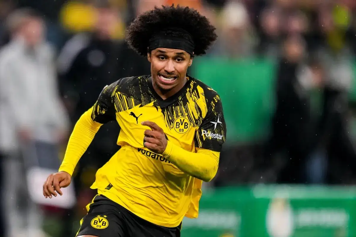 Karim Adeyemi w żółtej koszulce BVB z czarnymi wzorami, z afro i opaską na głowie, biegnie po boisku.