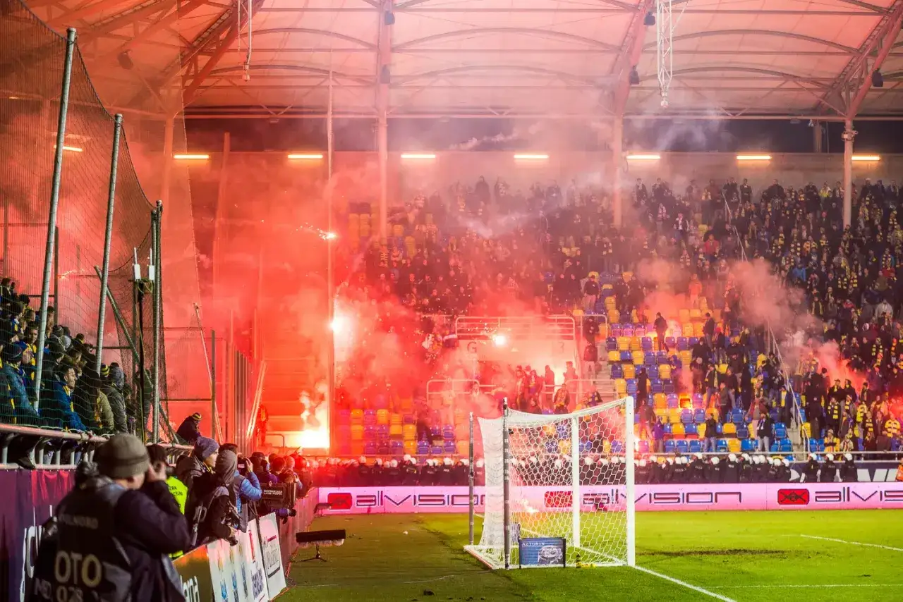 Gorąca atmosfera na stadionie podczas meczu. Kibice odpalają race, tworząc czerwony dym. To właśnie takie emocje towarzyszą meczom, które dla wielu są jak derby.