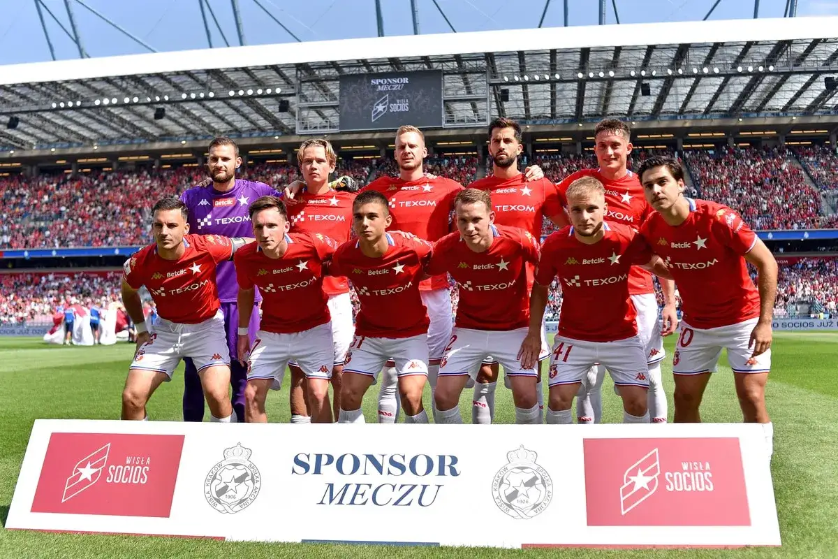 Piłkarze Wisły Kraków w czerwonych strojach, gotowi do meczu. Kibice na trybunach i baner 